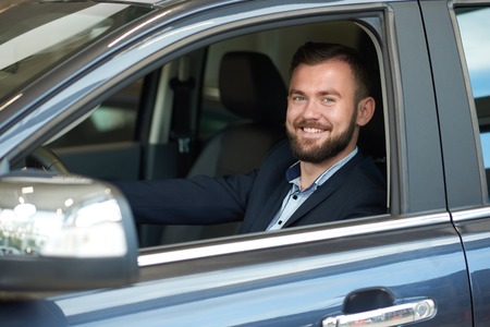 Smiling man sitting in car cabin, looking at camera and smiling. Bearded man holding hand on steering wheel, testing car before purchasing and buying. Customer observing dark blue automobile.の写真素材