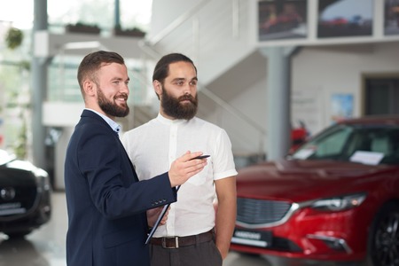 Bearded handsome men standing in car center and observing automobiles. Manager smiiling, holding folder and showing to customer cars. Brutal man in white shirt looking away and choosing.の写真素材