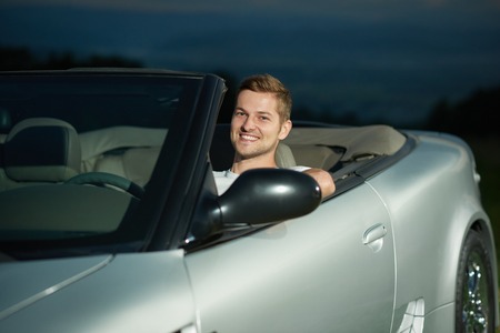 Handsome man sitting on drivers seat of silver cabriolet and posing. Male driver looking at camera...