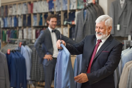 Handsome senior with grey hair choosing elegant clothes in store. Good looking man in black costume costume holding blue shirt on holder in boutique. Young attractive brunette standing on background.の写真素材