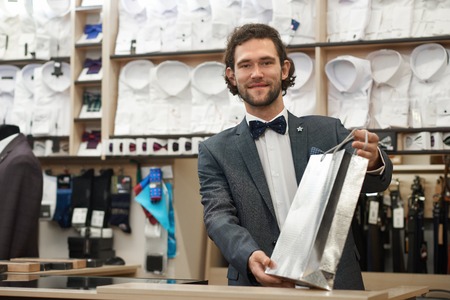 Handsome young man with beard holding silver package. Brunette attractive client buying present in boutique. Satisfied customer after shopping in male store with purchase.の写真素材