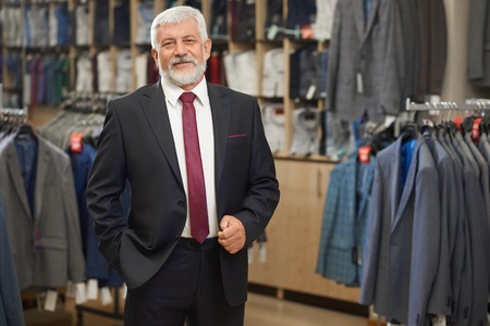 Old elegant man in grey costume with bow tie and white shirt posing at camera. Handsome man with beard smiling, holding hand in pocket. Shelves with shirts on background.の写真素材