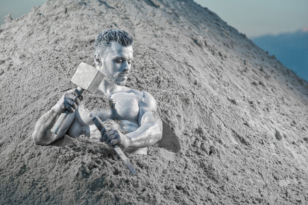 Handsome young man with beard lying on grey sand and thoughtfully looking on side. Mythical Atlas stuck among sand and saving himself with tools. Strong person creating his body.の写真素材
