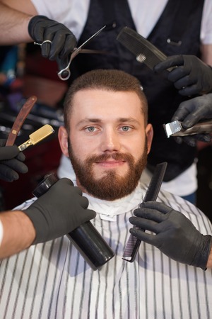 Portrait of smiling young client sitting in chair in barber shop and looking at camera while hands of male hairdressers keeping tools around. Several barbers styling hair and beard of customer.の写真素材