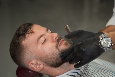 Closeup of handsome male client lying on chair with closed eyes during shaving in barber shop. Hands of professional barber in black gloves keeping straight razor and cutting beard.の写真素材