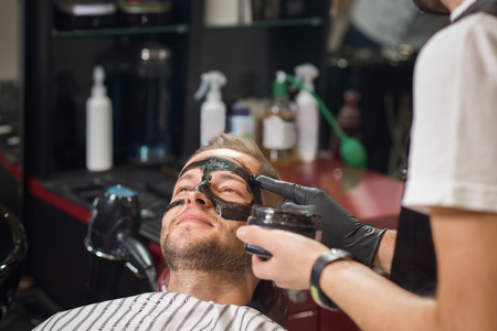 Young smiling male lying on chair in barber shop while barber in protective gloves putting black mask against black dots on face of client. Concept of cleaning procedure and face care.の写真素材