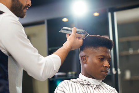 Side view of serious man sitting on chair and getting trendy haircut in barber shop. Barber in white shirt keeping clipper and correcting form of hair. Concept of hairstyling and trimming.の写真素材