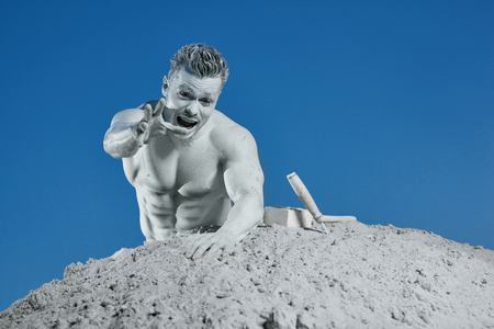 Man among grey sand screaming and showing by hand at camera. Atlas among sand saving himself with tools. Strong person creating his body with face expression and emotion. Blue sky background.の写真素材