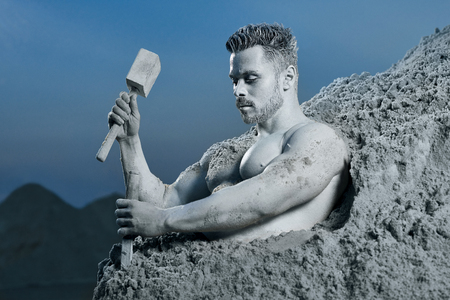 Close up of courage and handsome Atlas creating his perfect body from rock. Strong well build man with hammer in right hand and chisel in left looking down. Blue sky on background.の写真素材