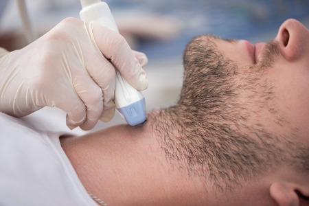Crop of male patient with beard lying in medical office on consultation. Specialist examining neck of man. Doctor in white medical gown holding devise and doing ultrasound diagnostics.の写真素材