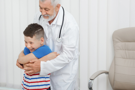 Happy kid on consultation with old doctor wearing in white medical gown. Therapist showing to boy exercises, holding his hands. Patient and physician smiling and looking away.の写真素材
