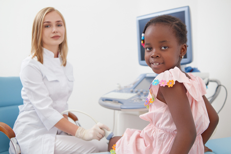 Side view of little cute afro girl sitting on bed and looking at camera during examines knee joints in hospital. Young female specialist, wearing in white medical gown, working with child at office.の写真素材