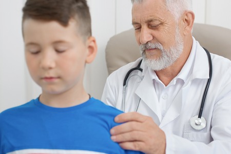 Old doctor with grey hair looking down at boys back and holding his shoulder. Kid standing with closed eyes. Professional therapist in white medical uniform having stethoscope on neck.の写真素材