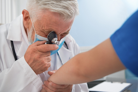 Professional therapist holding special tool near eye for diagnosis disease of skin. Doctor with grey hair wearing in medical uniform and mask. Physician examining hand of patient.の写真素材