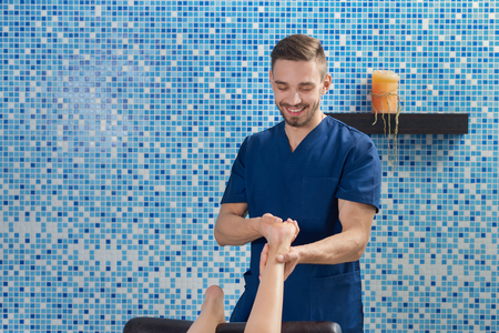 Front view of handsome male doctor in blue uniform smiling and talking while doing massage of feet in spa cosmetology salon. Masseur in process of working. Concept of treatment and care. Front view of handsome male doctor in blue uniform smiling and talking while doing massage of feet in spa cosmetology salon. Masseur in process of working. Concept of treatment and care.