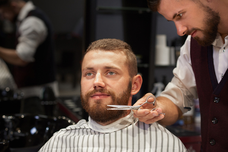 Smiling man sitting in chair in barber shop, looking away and getting cutting of beard. Qualified barber in white shirt and waistcoat standing near and styling male client with scissors.の写真素材