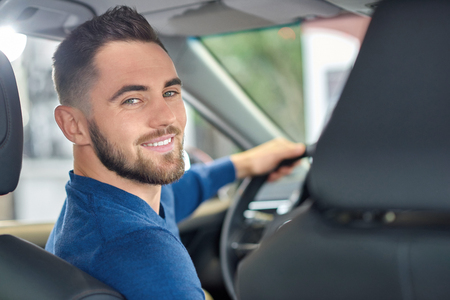 Portrait of hansome brunette man with beard looking at back. Young happy driver posing at camera and smiling. Attractive guy in blue sweater sitting at front seat and holding hand on stearing wheel.の写真素材