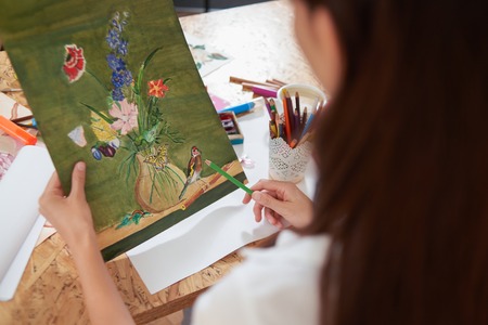 View from above of artist holding pencil and picture in hands. Illustration of beautiful flowers. Talented painter checking artwork. Table with painters supplies. Art school or studio.の写真素材