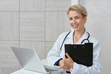 Portrait of seductive smiling female doctor in hospital. Blonde woman in white uniform looking at camera and smiling searching treatment. Concept of health care.の写真素材