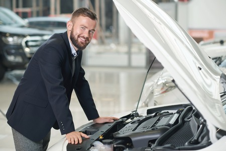 Customer of car dealership leaning on car with opened hood. Handsome man in dark blue jacket...