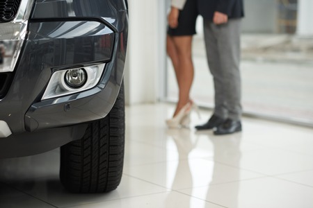 Headlight and tyre of expensive black car. Woman wearing high heels, skirt and man standing behind....