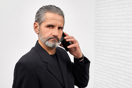 Serious businessman holding phone in hand near head, looking at camera. Mature bearded man wearing in black jacket posing in white studio. Concept of business and gadgets.の写真素材