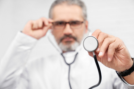 Close up of medical stethoscope. ENT doctor holding medical equipment, holding one hand on glasses. Physician looking at camera, posing in studio.の写真素材