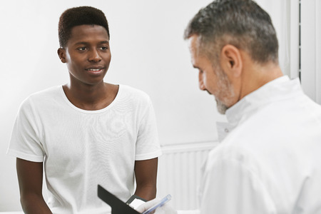 Handsome african wearing in white t shirt on consultation with his therapist. Patient looking at doctor, listening. Mature physician wearing in white medical uniform looking down.の写真素材