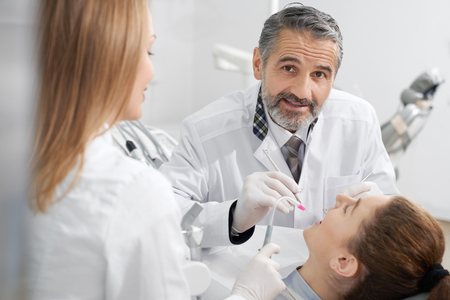 Woman with opened mouth lying in dentist chair. Handsome bearded dentist looking at camera, posing. Doctor with assistant healing teeth. Professional doctors using instruments.の写真素材