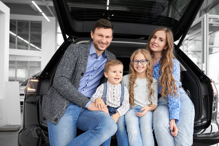 Cheerful and happy family posing together in car center showroom. Parents and children sitting in car trunk, luggage space of black automobile looking at camera and smiling.の写真素材