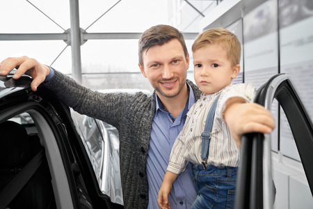 Handsome man holding hand on opened door of expensive black automobile. Cheerful father holding kid on hand. Parent and child smiling, looking at camera, posing in car dealership.の写真素材