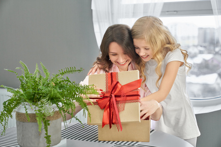 Joyful daughter and mom opening box with red bow and looking at it. Beautiful woman and cute child observing gift on table with houseplant. Girl with long, curly hair and brunette smiling.の写真素材