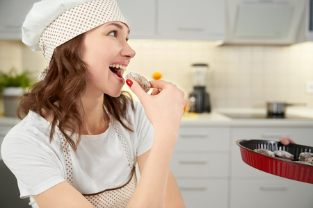 Beautiful, charming woman wearing in white chef hat and apron, looking away. Cheerful, pretty woman tasting, eating homemade, delicious cookie, holding biscuit in hand.の写真素材