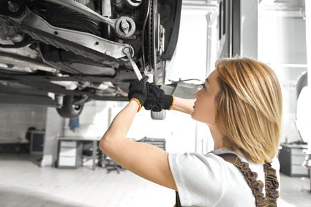 Blonde woman working as mechanic in autoservice, repairing car undercarriage, using wrench. Concentrated girl fixing vehicle in autoservice, with equipment. Automobile lifted on bridge.の写真素材