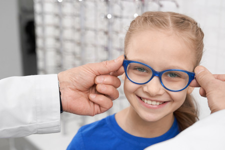 Close up of child in blue glasses. Male doctor ophthalmologist helping pretty girl to choose eyeglasses. Doctor and patient in ophthalmology clinic. Concept of health care.の写真素材