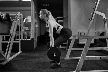 Side view of woman holding heavy barbell and working out in modern, equipped gym. Young, fit and athletic girl doing squats, wearing in black top, sportswear and sneakers.の写真素材