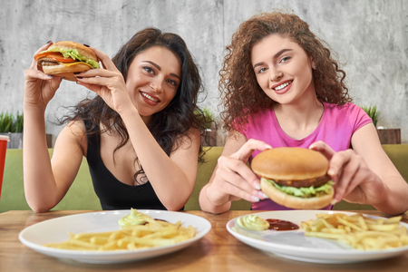 Two young, happy girls with beautiful curly hair sitting in cafe at table, looking at camera, smiling. Pretty brunette and blonde in colored t shirt holding hamburgers.の写真素材