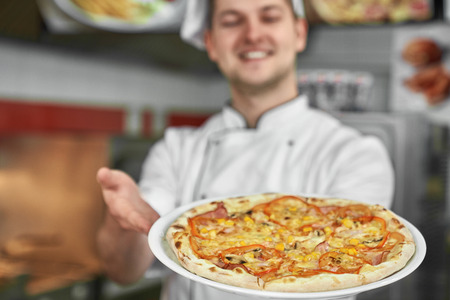Close up of pizza on white plate. Traditional italian cuisine. Positive, handsome chef in white uniform proposing delicious, tasty pizza in pizzeria. Concept of fast food, meal.の写真素材