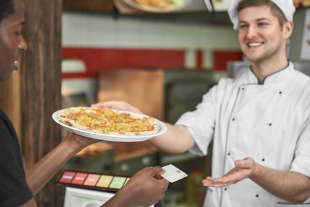 Young african holding delicious pizza on white plate and paying with card. Handsome, happy chef in white uniform smiling, giving dish. Concept of fast food and pizzeria.の写真素材