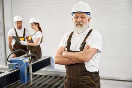 Professional elderly man posing with crossed hands on factory, Workers wearing in white helmets and t shirts, brown coveralls. Engineers standing near plasma laser cutter, talking.の写真素材