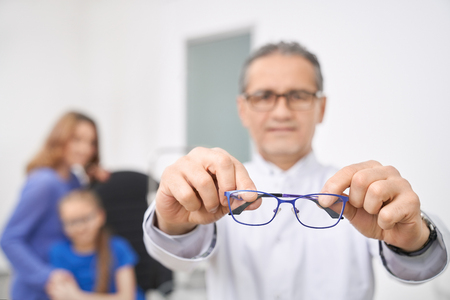 Close up of blue glasses. Doctor ophthalmologist holding, showing glasses. Medical worker in white coat looking at camera, standing in medical room of clinic. Patients on background.の写真素材