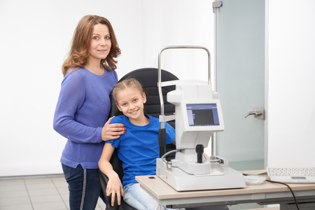 Pretty girl sitting in chair at table with modern slit lamp machine for examining eyesight. Mother and child posing in beautiful new optician office of ophthalmology clinic.の写真素材