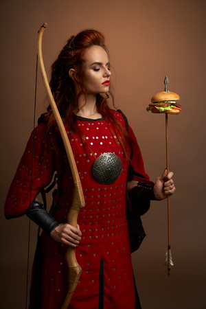 Medieval female warrior holding bow and arrow with hamburger, looking at tasty fast food burger. Gorgeous woman in red tunic with weapon posing and standing in studio.の写真素材