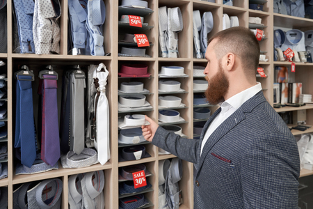 View from back of bearded male client standing near racks with clothes and choosing new shirt in clothes boutique. Young man wearing grey jacket looking at ties and shirt in shop.の写真素材