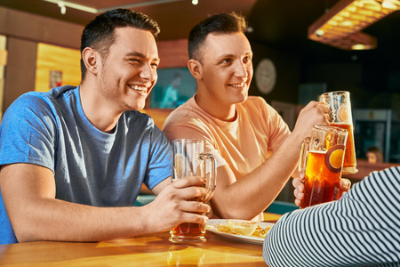 View from side of two happy men resting with friends in bar, drinking ale and laughing together....