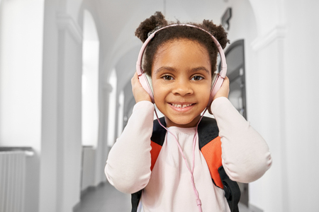 Positive, beautiful, cheerful schoolgirl posing with headphones in hallway of prrimary school. Pretty, smiling girl listening music in break between lessons.の写真素材