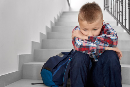 Disappointed, sad schoolboy of primary school with backpack sitting on stairwell. Handsome, depessed schoolchild wearing in checked shirt leaning head on hands.の写真素材