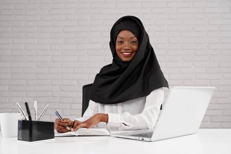 Beautiful, stylish african muslim woman wearing in black hijab posing at workplace, looking at camera. Female office worker sitting at table and working at laptop, writing in notebook.の写真素材
