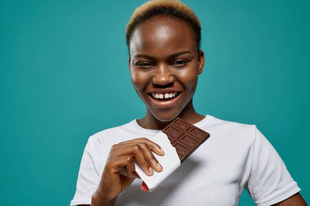 Happy, positive african woman holding bar of dark chocolate, smiling. Beautiful, charming, attractive girl in white t shirt posing on blue background, looking at camera.の写真素材
