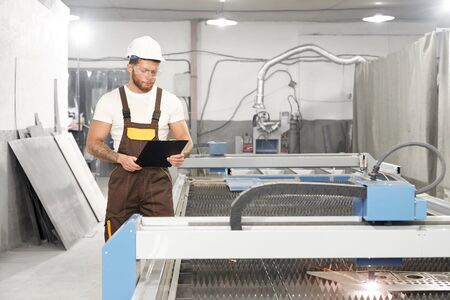 Front view of professional male mechanic standing near cnc and controlling process of plasma cutting...